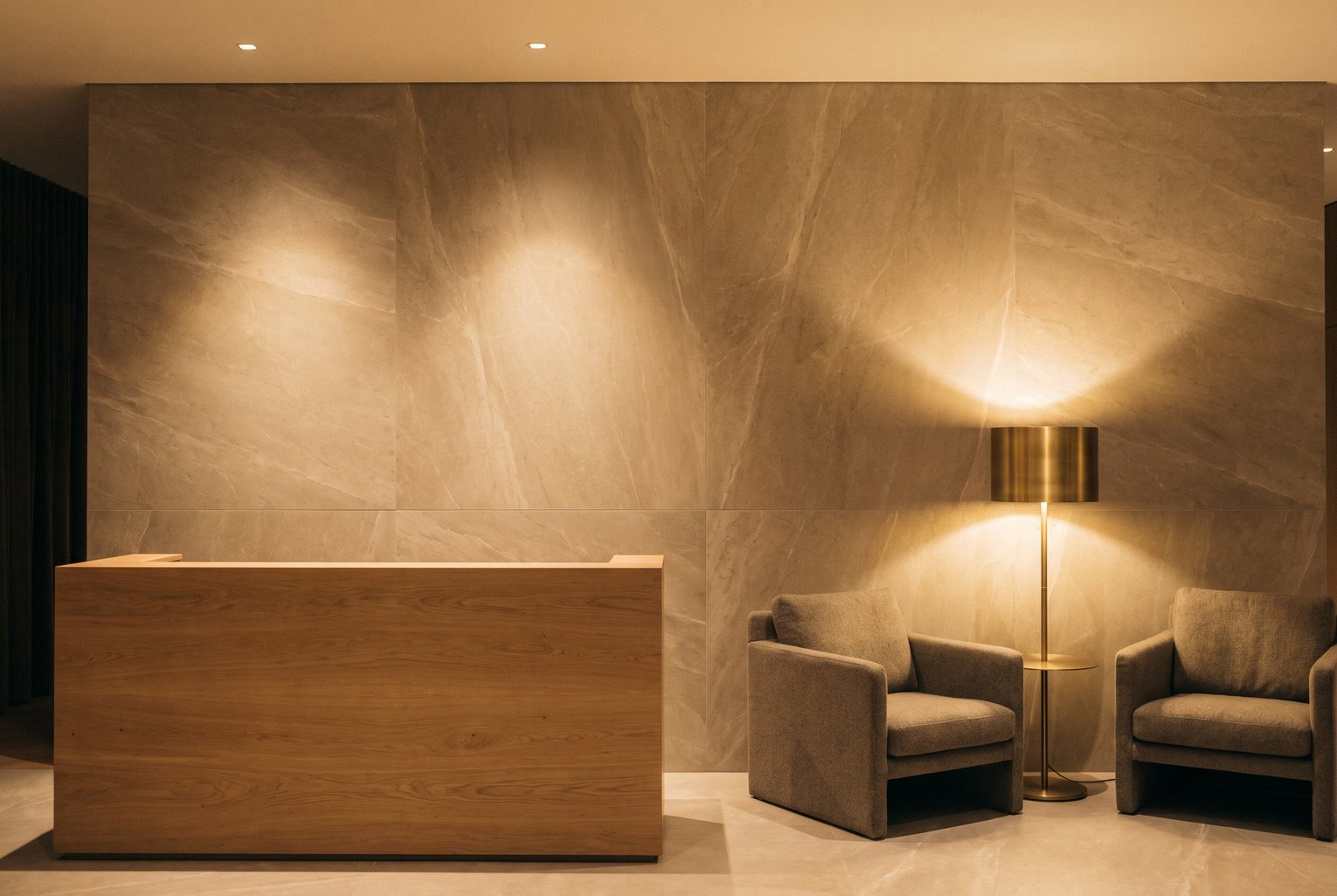 Réception d'hôtel éclairée de spots rasants, mur habillé de grands panneaux de pierre beige veinée derrière un comptoir en bois
