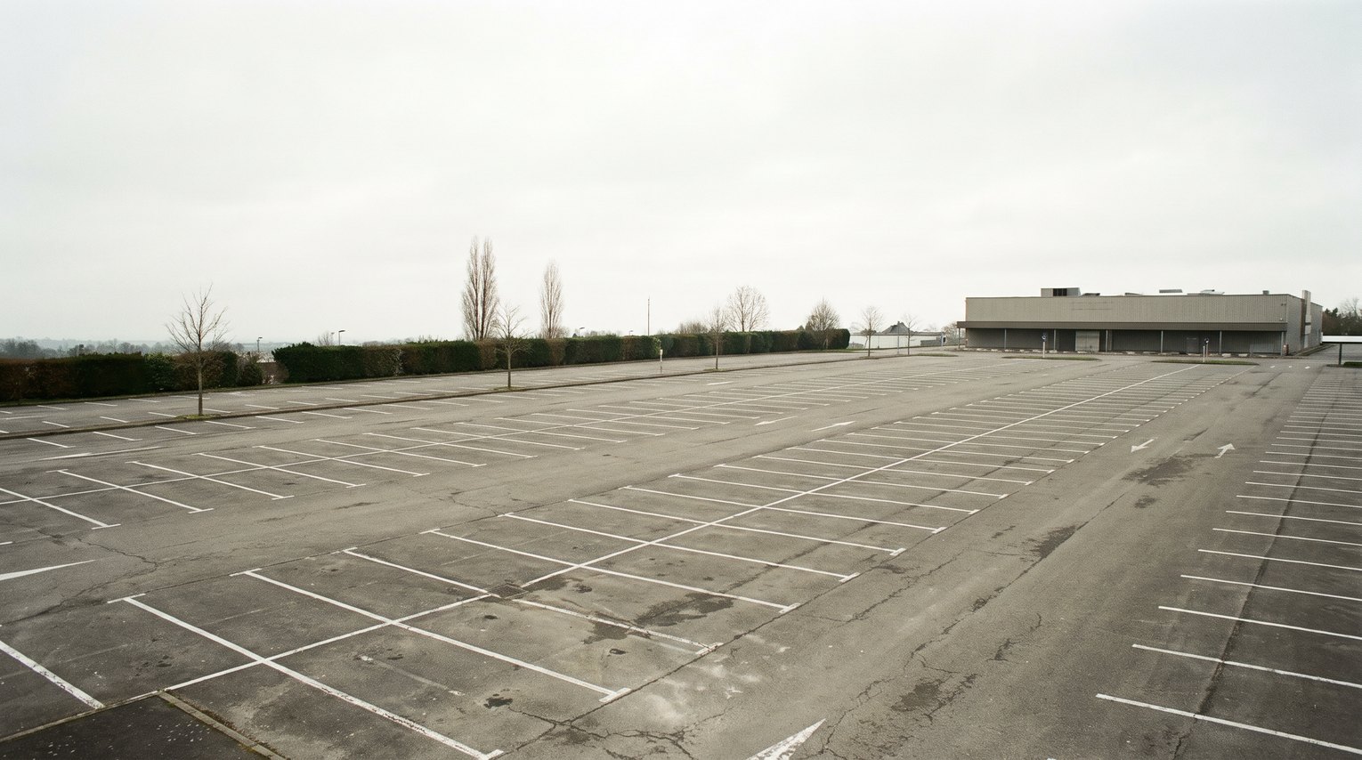 Grand parking extérieur vide et non couvert devant un bâtiment commercial, sous un ciel gris